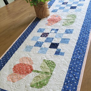May include: A rectangular quilted table runner featuring a white background with alternating blocks of blue patchwork squares and floral motifs in shades of coral and green. The runner is bordered by a dark blue fabric with a white floral pattern.