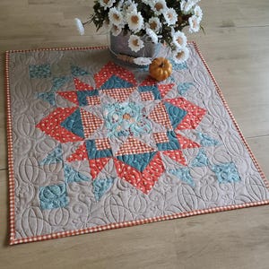 May include: A square quilted table topper featuring a central star pattern in shades of orange, teal, and white. The fabric has a beige background with intricate pumpkin-shaped machine quilting and a small orange and white checkered border trim.