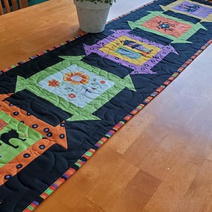 May include: A black quilted table runner with orange, green, and purple fabric squares featuring Halloween-themed designs. The squares are arranged in a row with a black border and a black and orange trim.