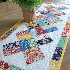 May include: A rectangular quilted table runner featuring a patchwork design of various floral and geometric fabric squares in red, blue, yellow, and green. The runner has a white background with intricate swirl stitching and a solid yellow border.