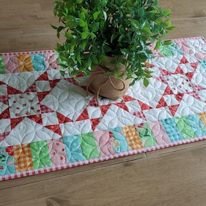 May include: A colorful quilted table runner with a star pattern in the center. The runner features a patchwork design with red, white, pink, green, and orange fabrics. A small potted plant sits on top of the runner.