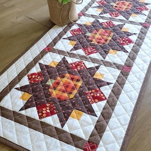 May include: A rectangular quilted table runner featuring three large star-shaped motifs in shades of brown, orange, red, and yellow. The runner has a cream-colored background with a diamond-stitched pattern and a solid brown fabric border.