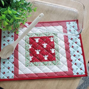 May include: A quilted, square hot pad with a red border and a central design featuring white angels on a red background. A wooden spoon and a glass baking dish rest on the pad, next to a green plant.