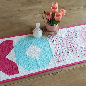 May include: A quilted table runner with a pink and white design. The runner features the letters "XO" in pink and turquoise, with a floral pattern and a pink border. A small bird figurine and a pot of tulips are on the runner.