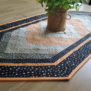 May include: A hexagonal quilted table runner featuring a geometric pattern in shades of orange, gray, and black. The fabric displays a repeating cross-stitch motif on the black borders and intricate pumpkin-shaped machine quilting throughout the center.