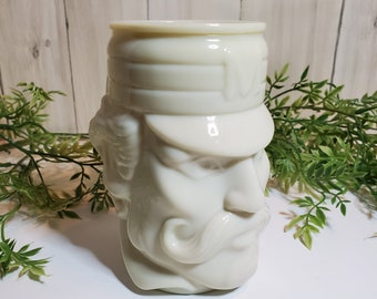 May include: A white ceramic mug shaped like a man's head with a mustache and a hat. The mug has a detailed face and is set against a white and wood background with green foliage.