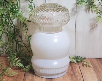 May include: A vintage white glass lamp with a clear, textured top. The lamp has a rounded base and a decorative, patterned top. The lamp is displayed on a wooden surface with green foliage in the background.