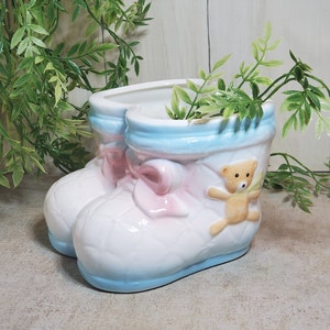 May include: A white ceramic baby boot planter with a quilted pattern, light blue trim, and a pink bow. A small tan teddy bear is attached to the side. The planter is filled with green leafy plants.