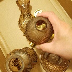 May include: An assortment of amber-colored glass perfume bottles and components. The bottles have textured, diamond-patterned surfaces. A hand holds a round bottle with a gold-colored metal base. The items are arranged in a cardboard box.