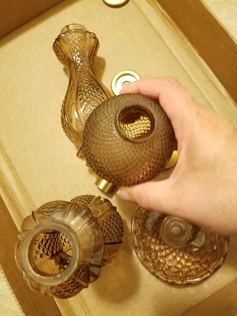 May include: An assortment of amber-colored glass perfume bottles and components. The bottles have textured, diamond-patterned surfaces. A hand holds a round bottle with a gold-colored metal base. The items are arranged in a cardboard box.