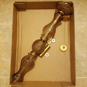 May include: A disassembled glass lamp with a brown tint, featuring a decorative pattern and gold-colored metal accents. The lamp is in a cardboard box, along with small metal pieces.
