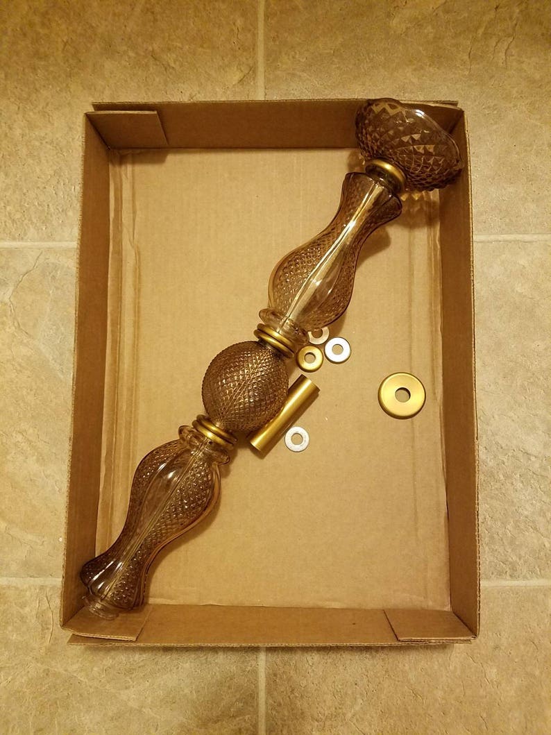 May include: A disassembled glass lamp with a brown tint, featuring a decorative pattern and gold-colored metal accents. The lamp is in a cardboard box, along with small metal pieces.