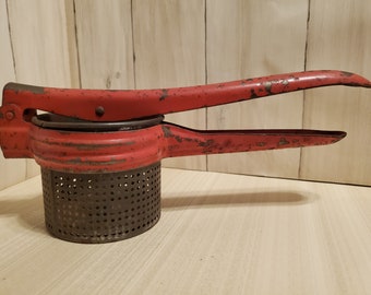May include: Vintage red potato ricer with a perforated metal base and long handles. The kitchen tool shows signs of wear, with chipped paint and a rustic appearance. A classic utensil for mashing potatoes or other soft foods.