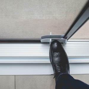 May include: A person's foot steps on a white foot pedal attached to a glass door. The foot pedal is designed to open the door without using hands.