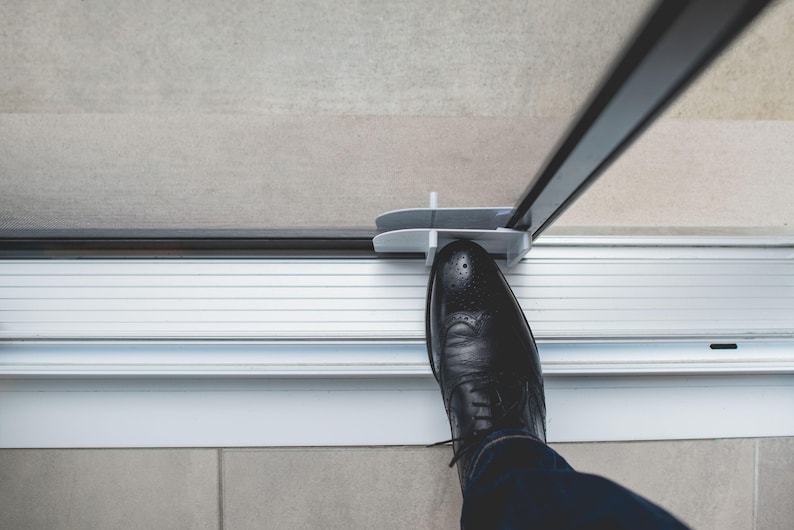 May include: A person's foot steps on a white foot pedal attached to a glass door. The foot pedal is designed to open the door without using hands.