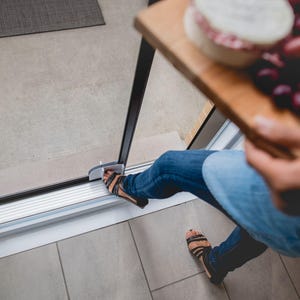 May include: A person wearing blue jeans and sandals steps through a sliding glass door. The person is holding a wooden tray with a round cheese and grapes.