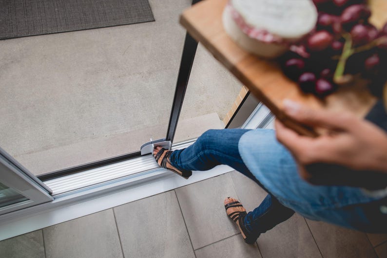 May include: A person wearing blue jeans and sandals steps through a sliding glass door. The person is holding a wooden tray with a round cheese and grapes.