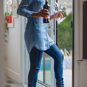 May include: A woman wearing a blue denim shirt and jeans stands by a sliding glass door, holding a bottle of wine and two wine glasses.