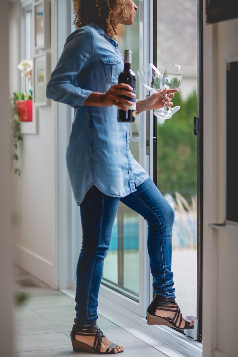 May include: A woman wearing a blue denim shirt and jeans stands by a sliding glass door, holding a bottle of wine and two wine glasses.
