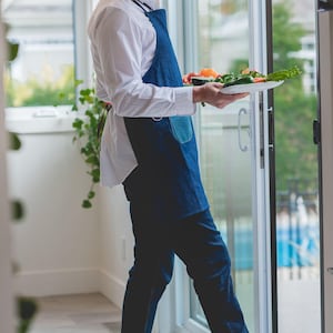 May include: A man wearing a blue denim apron and a white shirt is holding a plate of fresh vegetables. He is standing in a kitchen with a sliding glass door behind him.