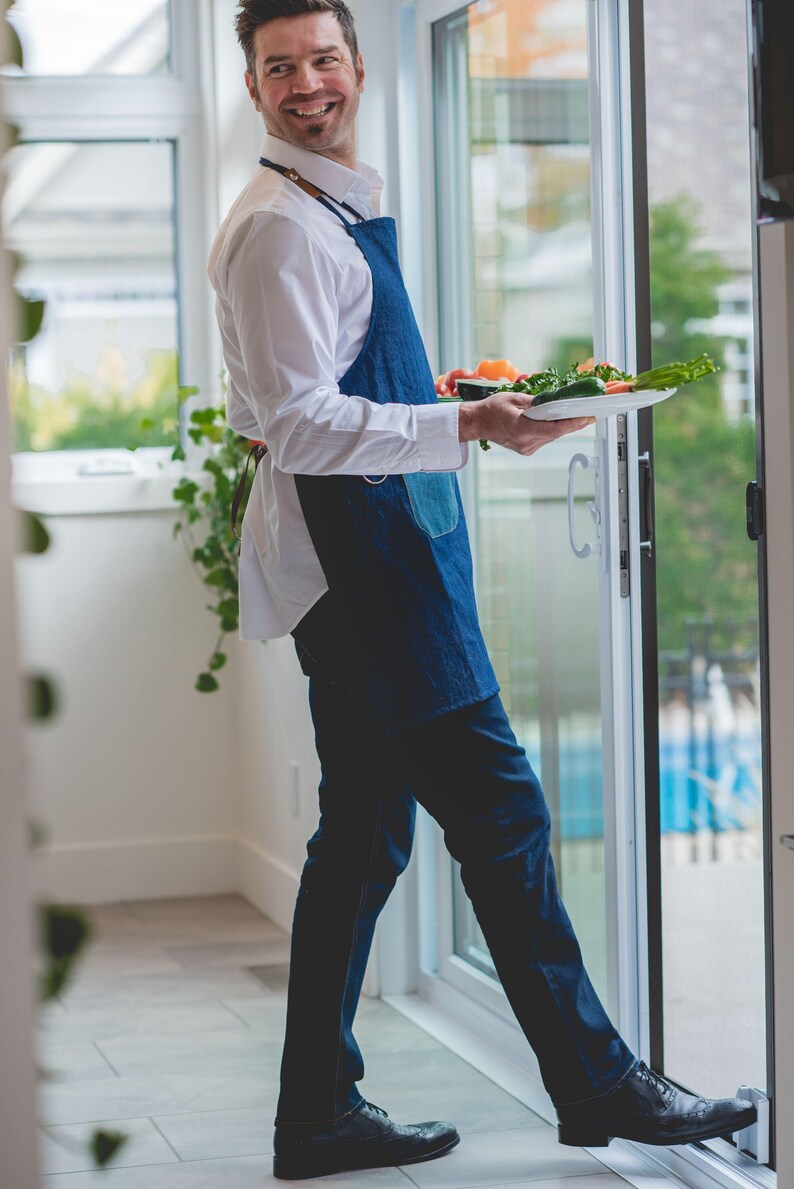 May include: A man wearing a blue denim apron and a white shirt is holding a plate of fresh vegetables. He is standing in a kitchen with a sliding glass door behind him.