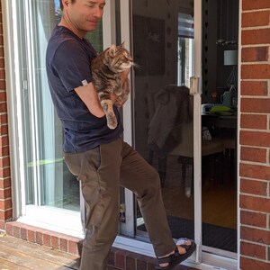 May include: A man wearing a blue shirt and brown trousers holds a tabby cat in his arms. He is standing in front of a sliding glass door with a screen.