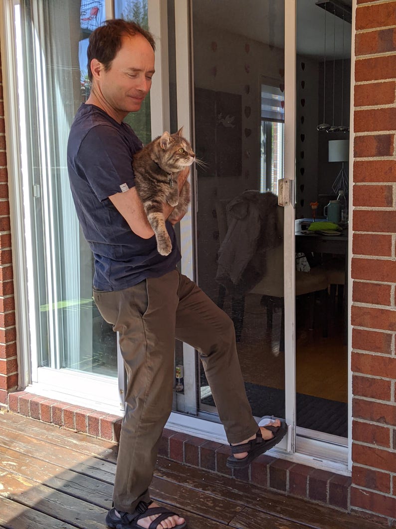 May include: A man wearing a blue shirt and brown trousers holds a tabby cat in his arms. He is standing in front of a sliding glass door with a screen.