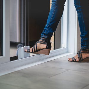 May include: A person wearing blue jeans and black sandals with a wedge heel is opening a sliding glass door. The door has a white frame and a clear glass panel.