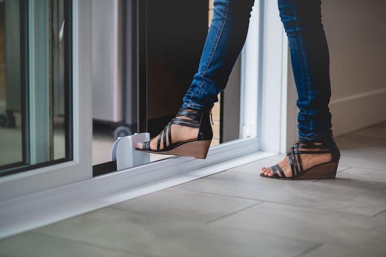 May include: A person wearing blue jeans and black sandals with a wedge heel is opening a sliding glass door. The door has a white frame and a clear glass panel.