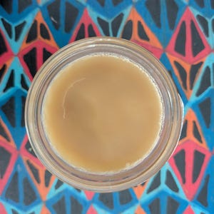 May include: A top-down view of a clear glass container filled with a light tan, opaque liquid. The container is positioned against a vibrant background featuring a repeating geometric pattern of black, bright pink, and light blue diamond shapes.