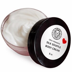 May include: A jar of Ultra Aromatics Silk Souffle Body Cream. The clear jar is open, revealing a swirl of white cream. The black lid is next to the jar, with the product name and a lion logo on a white label. The label also includes a red heart.
