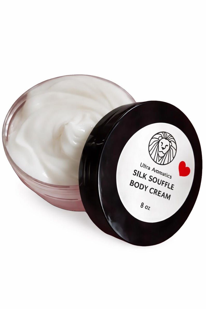 May include: A jar of Ultra Aromatics Silk Souffle Body Cream. The clear jar is open, revealing a swirl of white cream. The black lid is next to the jar, with the product name and a lion logo on a white label. The label also includes a red heart.