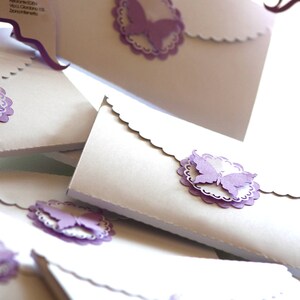 Puede incluir: Una pila de sobres blancos con bordes festoneados y adornos de mariposas moradas. Los sobres est&aacute;n decorados con un delicado patr&oacute;n de encaje.