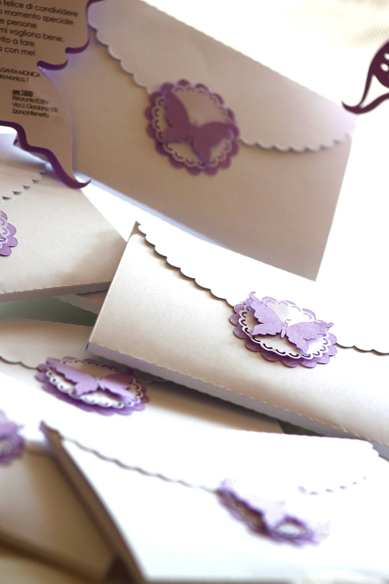 Puede incluir: Una pila de sobres blancos con bordes festoneados y adornos de mariposas moradas. Los sobres est&aacute;n decorados con un delicado patr&oacute;n de encaje.
