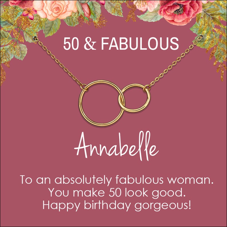 May include: A gold necklace with two interlocking circles, a pink card with the text "50 & FABULOUS" and the name "Annabelle" in white script. The card also includes the text "To an absolutely fabulous woman. You make 50 look good. Happy birthday gorgeous!"