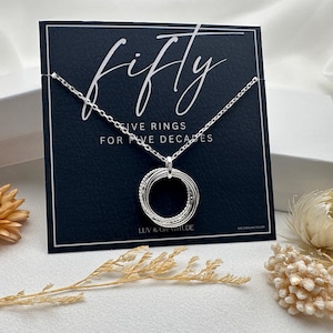 May include: A silver necklace with a pendant that says "fifty" and "five rings for five decades". The necklace is displayed on a black card with a white background.
