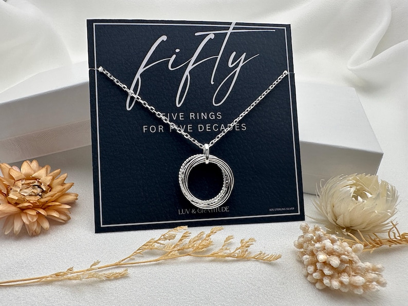 May include: A silver necklace with a pendant that says "fifty" and "five rings for five decades". The necklace is displayed on a black card with a white background.