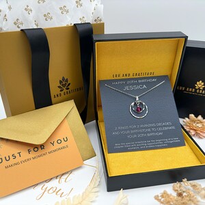 May include: A gold gift box with a black lid and a black and white necklace inside. The necklace has a silver chain and a pendant with two rings and a red gemstone. The box has a yellow interior and a card that says "LUV AND GRATITUDE" and "HAPPY 20TH BIRTHDAY JESSICA".