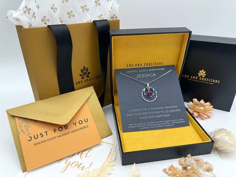 May include: A gold gift box with a black lid and a black and white necklace inside. The necklace has a silver chain and a pendant with two rings and a red gemstone. The box has a yellow interior and a card that says "LUV AND GRATITUDE" and "HAPPY 20TH BIRTHDAY JESSICA".