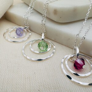 May include: Three silver necklaces with delicate chain links and pendant charms. Each pendant features a single crystal stone in a circle design. The stones are purple, green, and red.