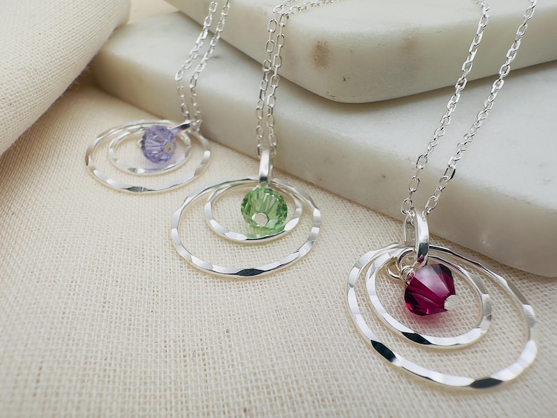 May include: Three silver necklaces with delicate chain links and pendant charms. Each pendant features a single crystal stone in a circle design. The stones are purple, green, and red.