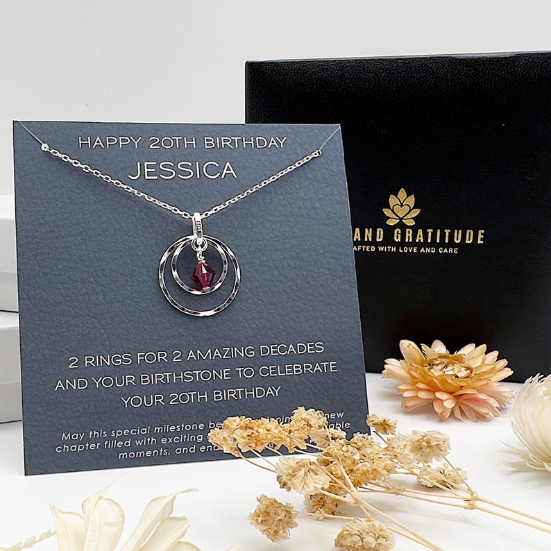 May include: A silver necklace with two rings and a red gemstone pendant. The necklace is displayed on a gray card with the text "Happy 20th Birthday Jessica" and "2 rings for 2 amazing decades and your birthstone to celebrate your 20th birthday".