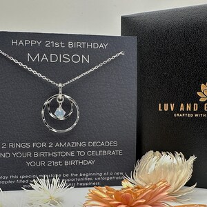 May include: A silver necklace with a double ring pendant and a clear crystal charm. The necklace is displayed on a black card with the text "Happy 21st Birthday Madison" and "2 rings for 2 amazing decades and your birthstone to celebrate your 21st birthday".