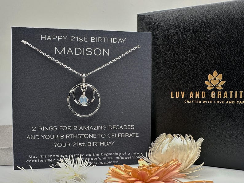 May include: A silver necklace with a double ring pendant and a clear crystal charm. The necklace is displayed on a black card with the text "Happy 21st Birthday Madison" and "2 rings for 2 amazing decades and your birthstone to celebrate your 21st birthday".