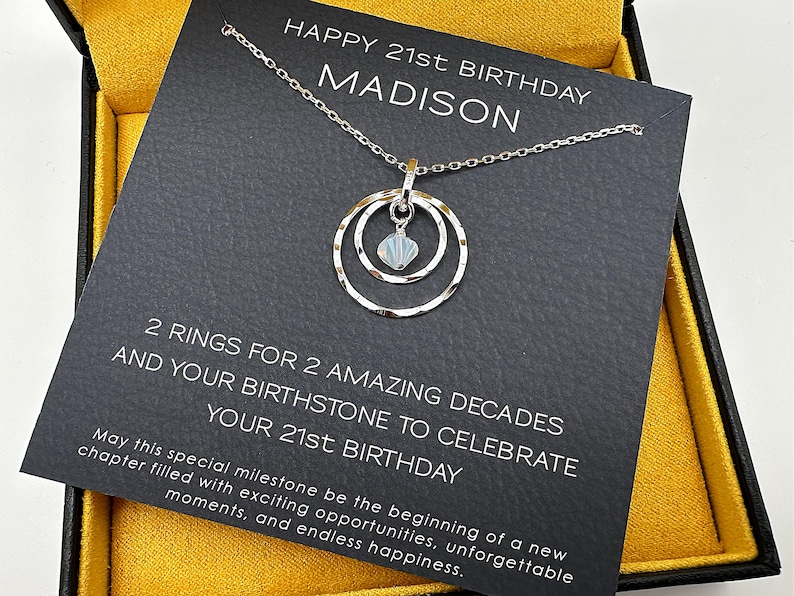 May include: A black card with the text "HAPPY 21st BIRTHDAY MADISON" and "2 RINGS FOR 2 AMAZING DECADES AND YOUR BIRTHSTONE TO CELEBRATE YOUR 21st BIRTHDAY". A silver necklace with a double circle pendant and a small white stone hangs from the top of the card.