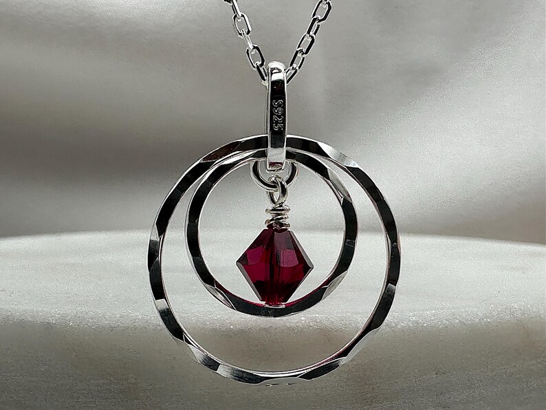 May include: A silver necklace with a pendant featuring two concentric circles and a dark red gemstone in the center. The pendant has the inscription "S925" on the back.