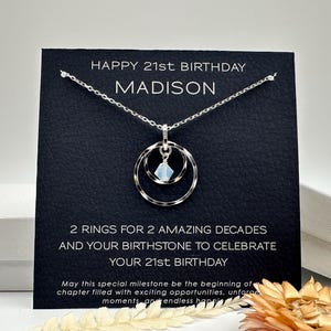 May include: A black card with the text "HAPPY 21st BIRTHDAY MADISON" and "2 RINGS FOR 2 AMAZING DECADES AND YOUR BIRTHSTONE TO CELEBRATE YOUR 21st BIRTHDAY". A silver necklace with a double ring pendant and a light blue gemstone hangs from the card.