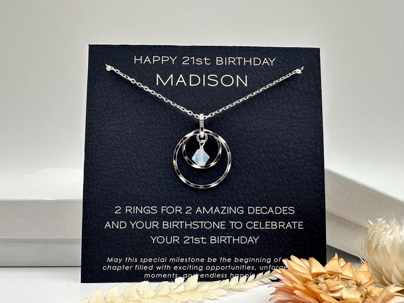 May include: A black card with the text "HAPPY 21st BIRTHDAY MADISON" and "2 RINGS FOR 2 AMAZING DECADES AND YOUR BIRTHSTONE TO CELEBRATE YOUR 21st BIRTHDAY". A silver necklace with a double ring pendant and a light blue gemstone hangs from the card.