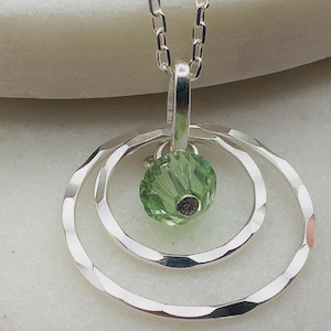 May include: A silver necklace with a pendant featuring three concentric circles and a green crystal in the centre.
