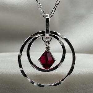 May include: A silver necklace with a pendant featuring two interlocking circles and a dark red crystal. The pendant is marked with "S925".